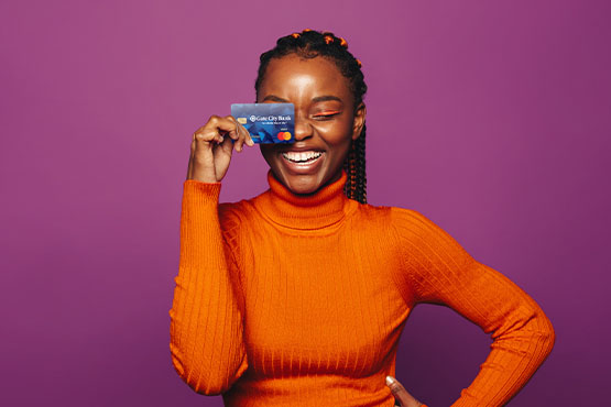 Close-up of a young woman in an orange turtleneck, smiling and holding her Gate City Bank debit card over one eye