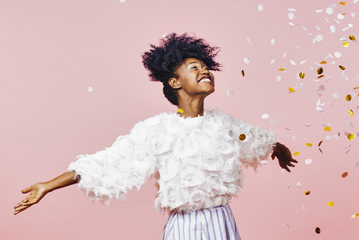 happy young woman looks up and holds arms out as confetti falls down around her