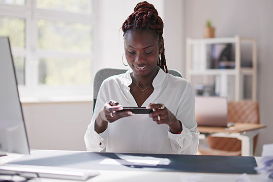 a happy woman sits at a desk and uses mobile deposit on her phone after reading Gate City Bank’s tips to prevent check fraud