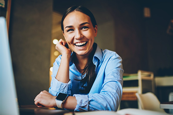 Smiling businesswoman with dimples, wearing a blue shirt and sitting by a laptop inside her small business in Fargo, ND