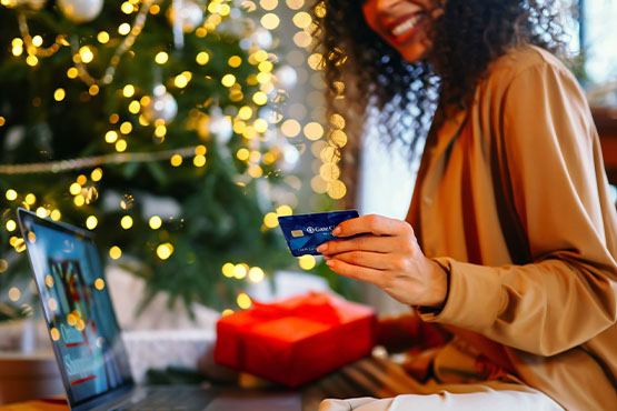 A young woman sits by a Christmas tree, using her debit card to save money while holiday shopping