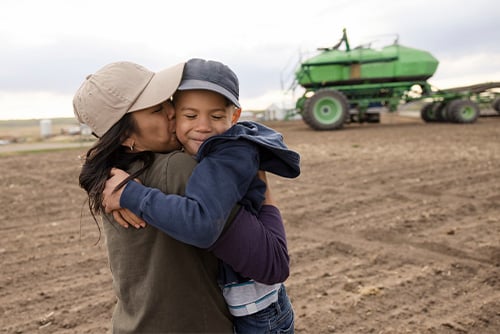 Smiling boy in a baseball cap, getting a bear hug from his mom on their farm outside Wahpeton, ND