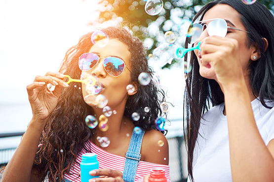 two young women wearing aviator sunglasses, blowing bubbles on a park bench in Fargo, ND