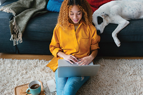 Young woman with curly hair, accessing her Preferred Perks Checking benefits on a laptop while sitting on living room floor