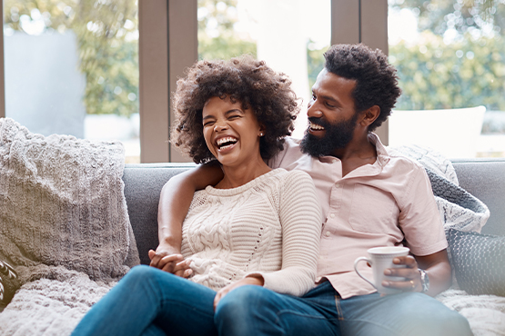 Carefree millennial couple cuddling on a gray couch, laughing after refinancing their mortgage with Gate City Bank in St. Cloud, MN