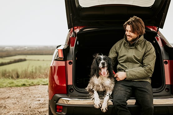 a man sits beside his dog under his car’s open hatch after reading an article on buying a new car vs used from Gate City Bank