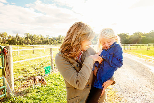 Smiling mom holds her young blonde daughter on her hip, standing on a gravel road on their farm outside Mayville, ND