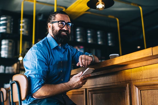 Small business owner with beard and glasses, sitting at his bar counter and banking on his phone in downtown Fargo, ND
