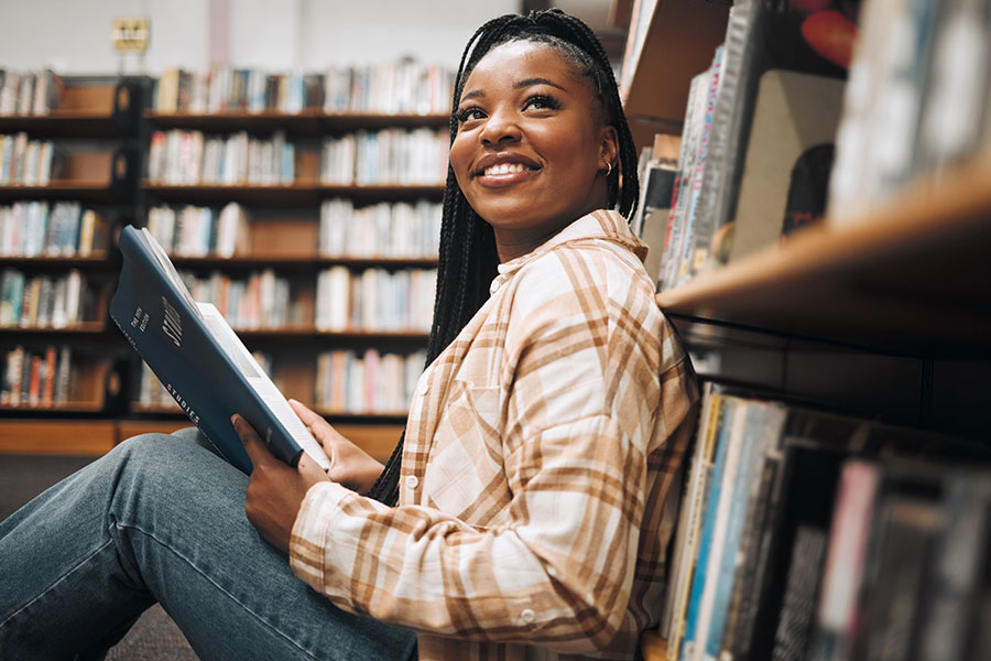 Young NDSU student wearing a plaid pink and tan shirt, smiling while sitting on the library floor with a book