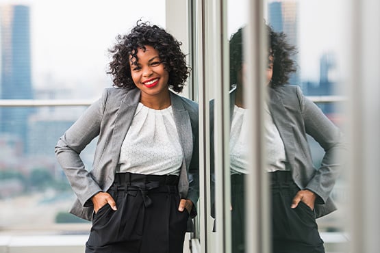 Smiling businesswoman with curly hair in a gray suit, standing on her office terrace with the Grand Forks, ND, skyline behind her