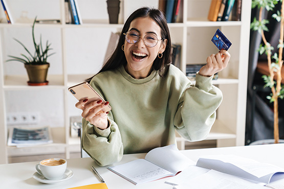 a young woman holding a phone and debit card smiles at her desk after learning about debit card benefits from Gate City Bank