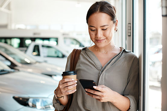 Young woman drinking coffee at a car dealership, using a calculator on her phone to compare rates and rebates
