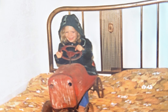 A young Kim Settel sits on her vintage red pedal car, which is parked on top of a bed