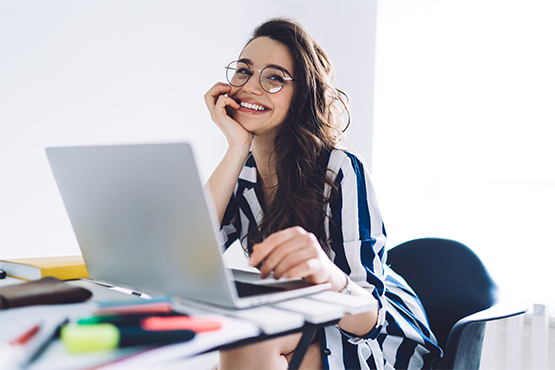 a woman wearing glasses smiles and glances to the side while using her laptop to learn about Gate City Bank’s IRA options