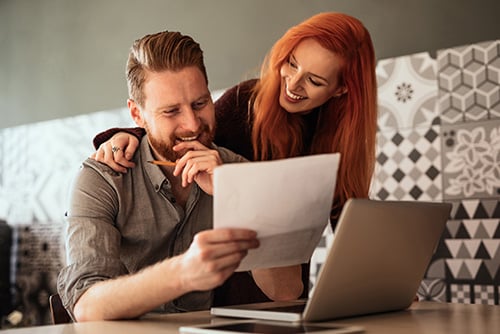 happy young couple looking at a laptop on their kitchen table, discussing their wealth management and financial planning options