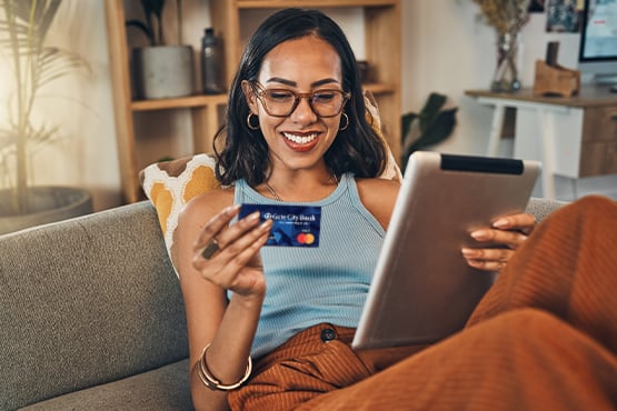 a happy woman holding a credit card & tablet sits on her couch after learning 5 ways to consolidate debt from Gate City Bank
